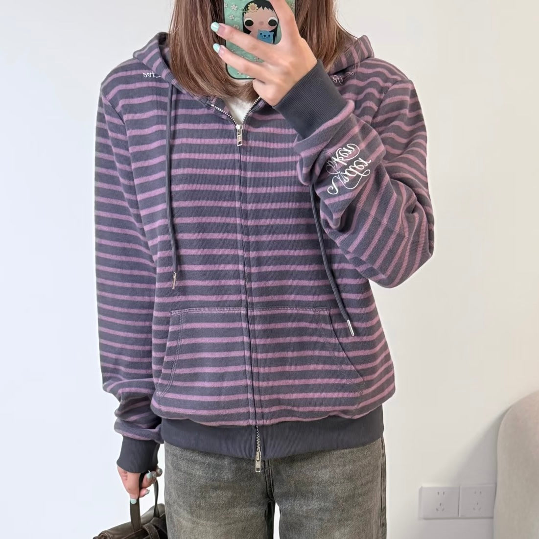 A femboy takes a mirror selfie wearing a purple and pink striped zip-up hoodie with dark gray ribbed cuffs, hem, and drawstrings, paired with gray jeans.