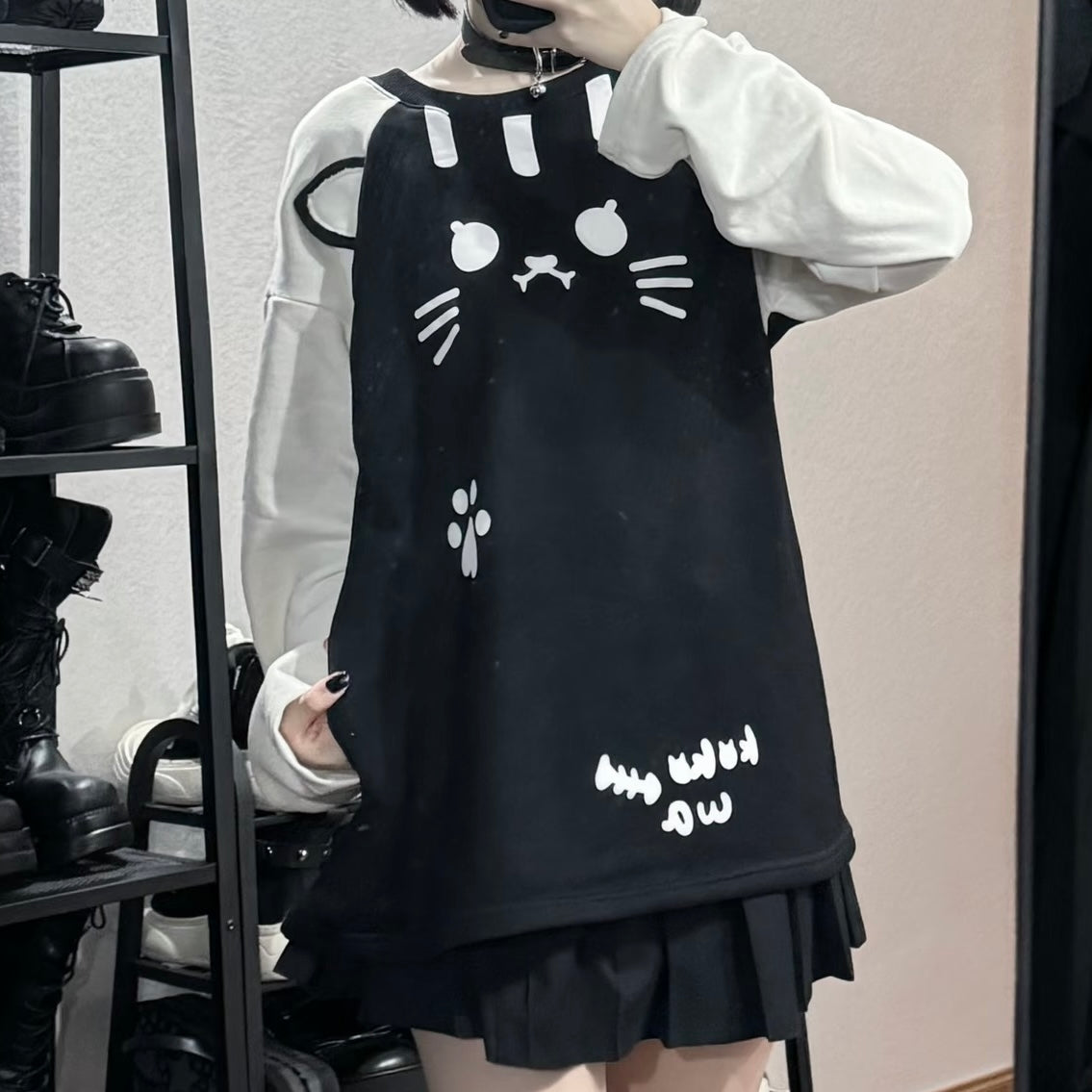 A femboy wearing a black cat-face print mini dress over a white long-sleeve shirt, standing in front of a mirror in what appears to be a boutique.