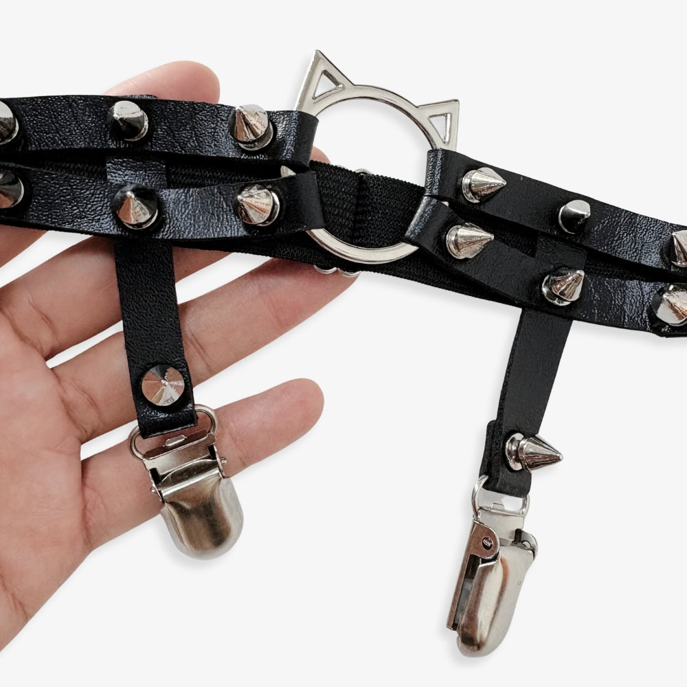 Black leather thigh garters with spikes and clips held by a hand on a white background