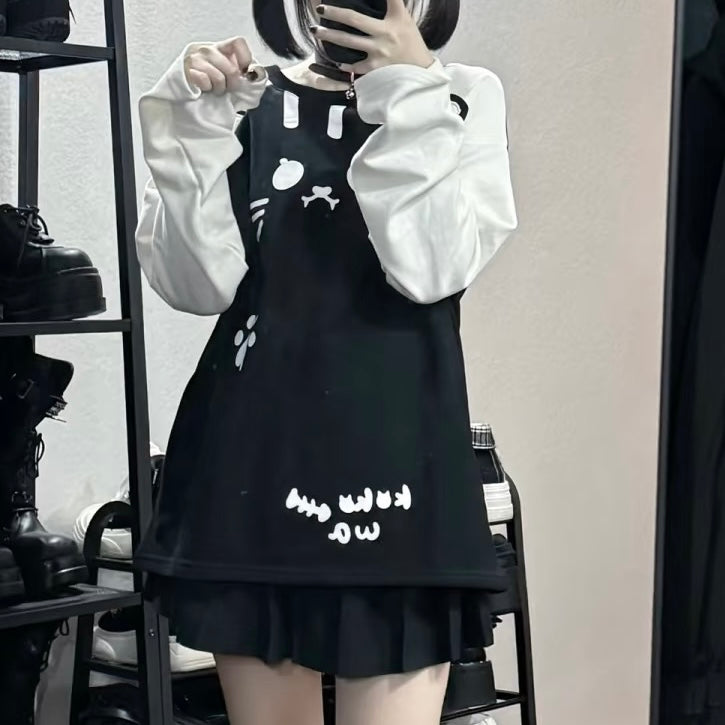 A femboy takes a mirror selfie wearing a black sleeveless pinafore-style dress with white cartoon graphics and text, layered over a white long-sleeve top. The setting appears to be a boutique or room with shelves and shoes visible in the background.