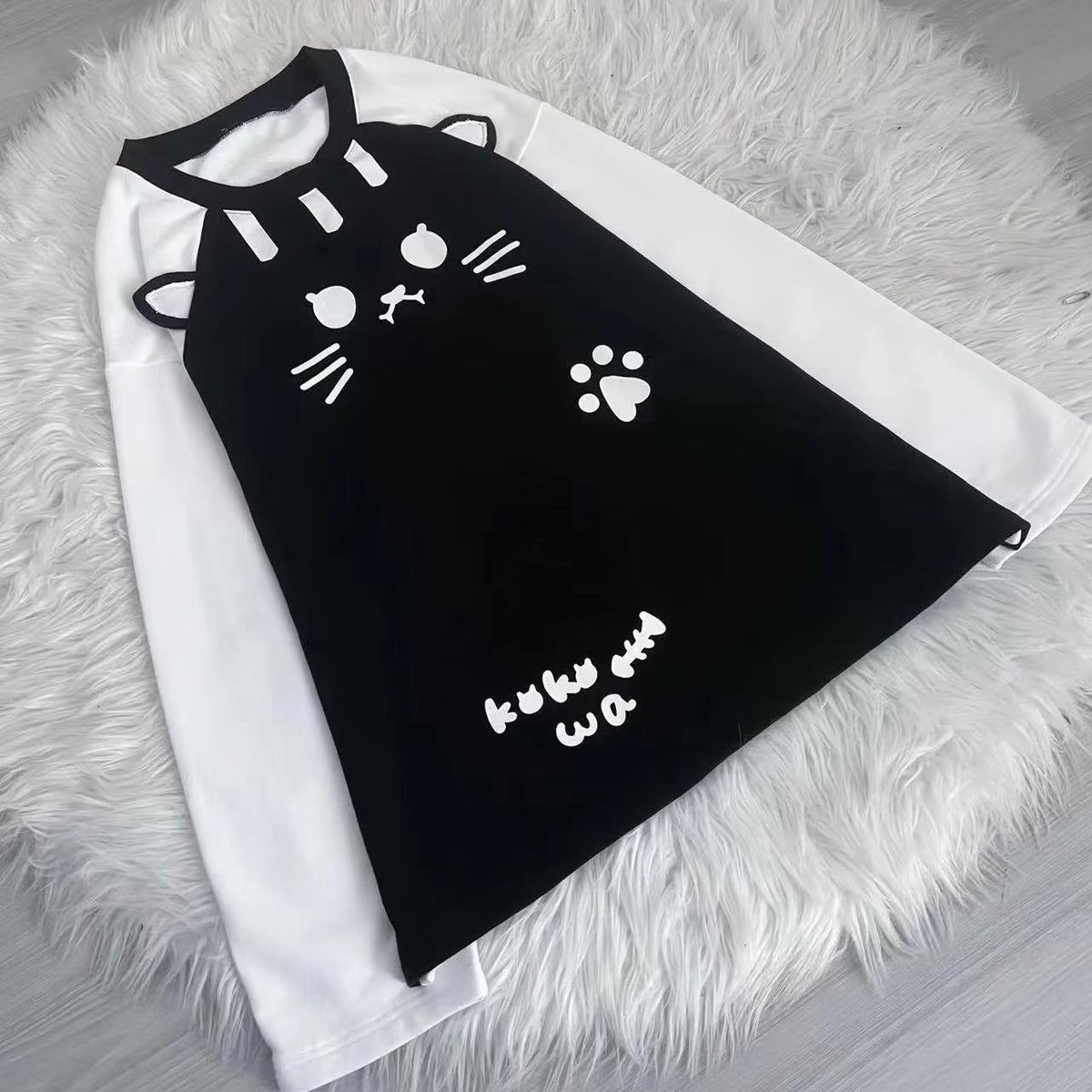 A black and white raglan long-sleeve shirt featuring a cute cat face, whiskers, paw print, and 'kuku wa' text on the front, laid flat on a white faux fur rug.
