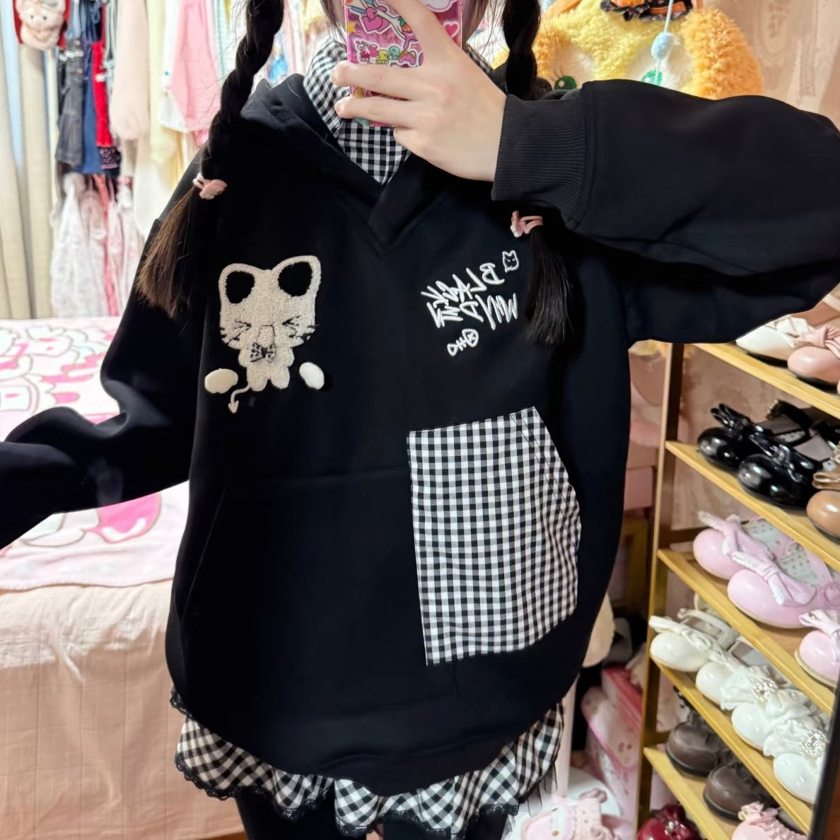 Person wearing a black hoodie with white text and graphics, standing in a room with shelves of shoes and clothes.