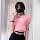 Person wearing a pink crop top with a graphic design and black pants, standing against a light curtain background.