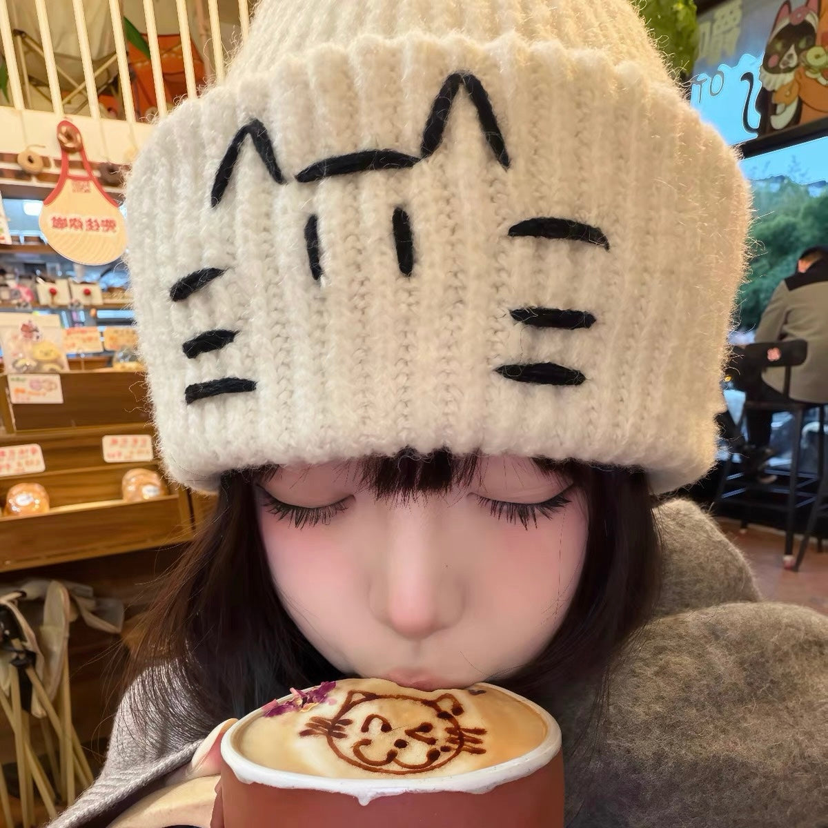 Person wearing a cat-themed knit hat, drinking a latte with a cat design.