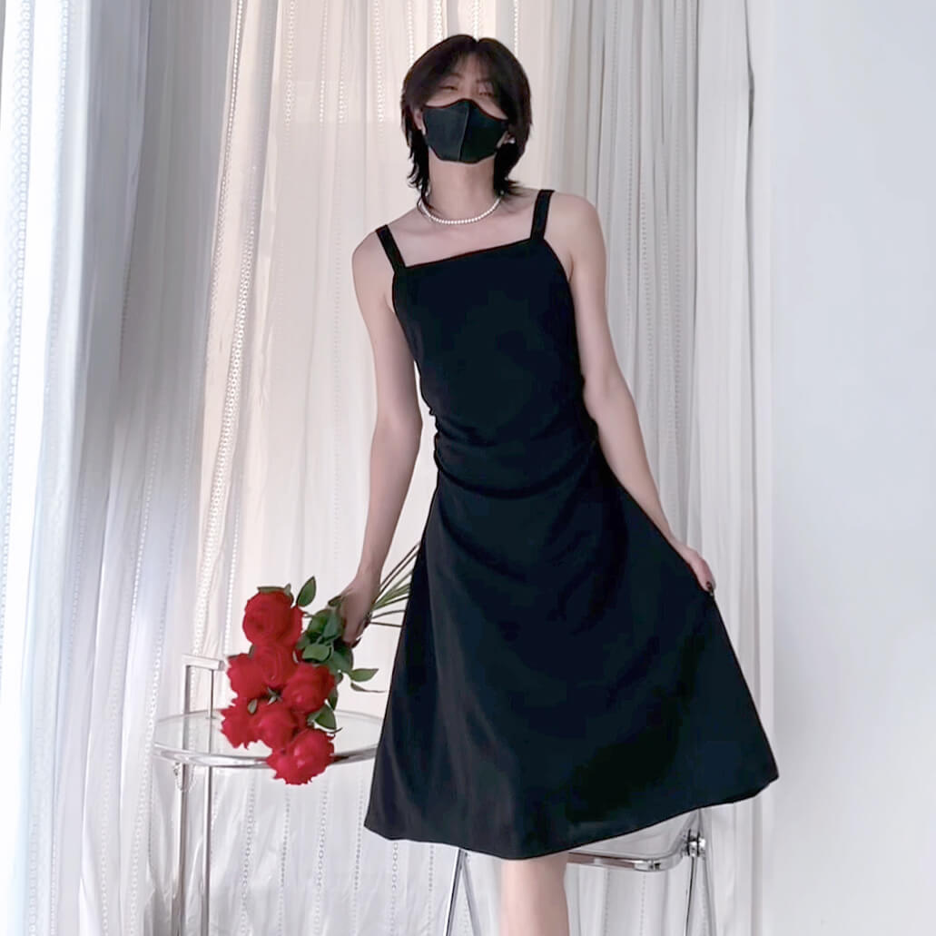 Femboy in a black dress holding red flowers against a white curtain background