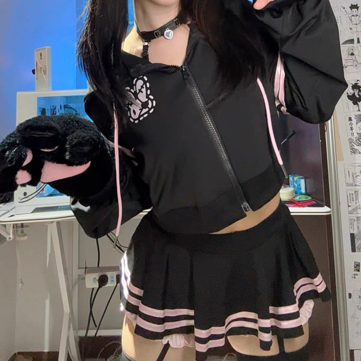 A femboy wearing a black crop hoodie and mico skirt set, holding a black plush toy in a room setting.