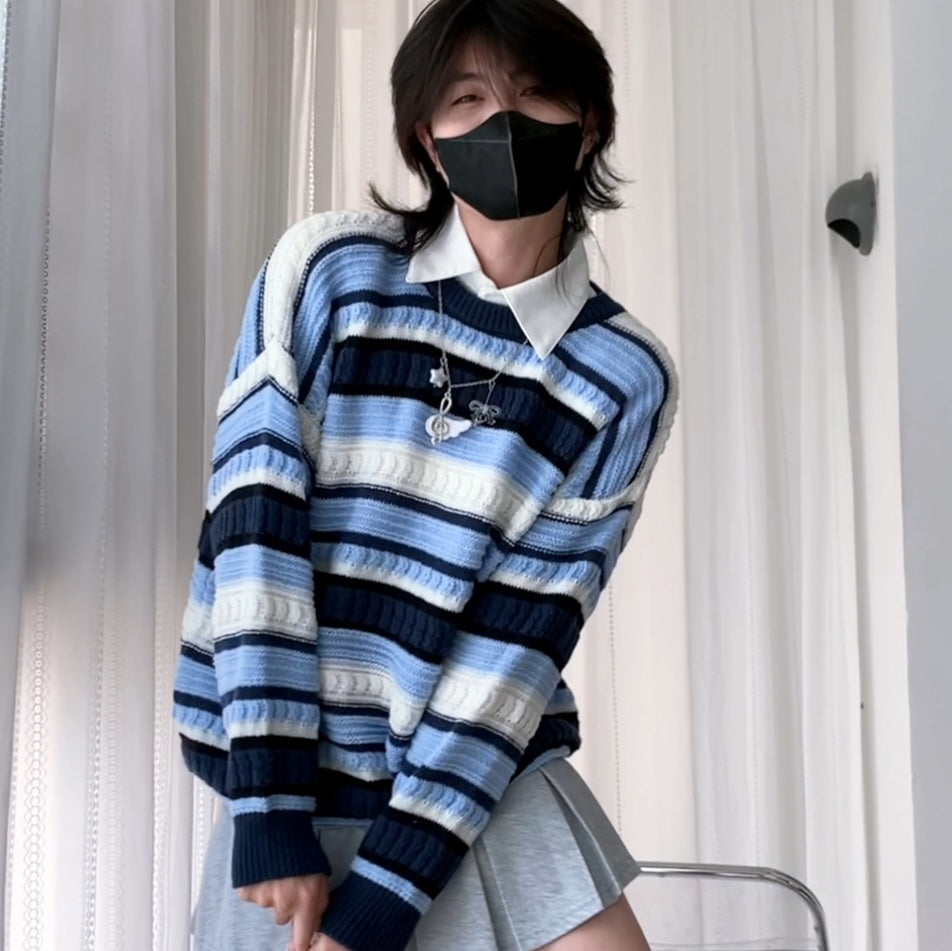 A femboy wearing a blue, navy, and white striped knit sweater over a white collared shirt, paired with a gray skirt, standing in front of white curtains.