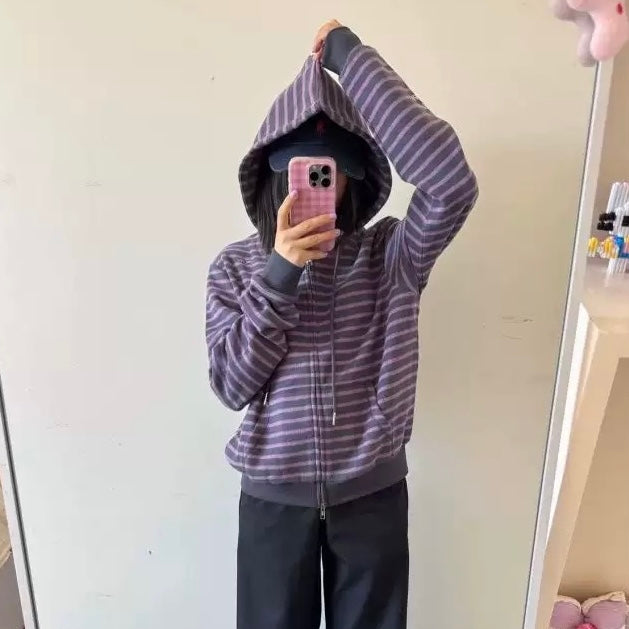 A femboy taking a mirror selfie wearing a purple and gray striped zip-up hoodie, black pants, and a black cap.
