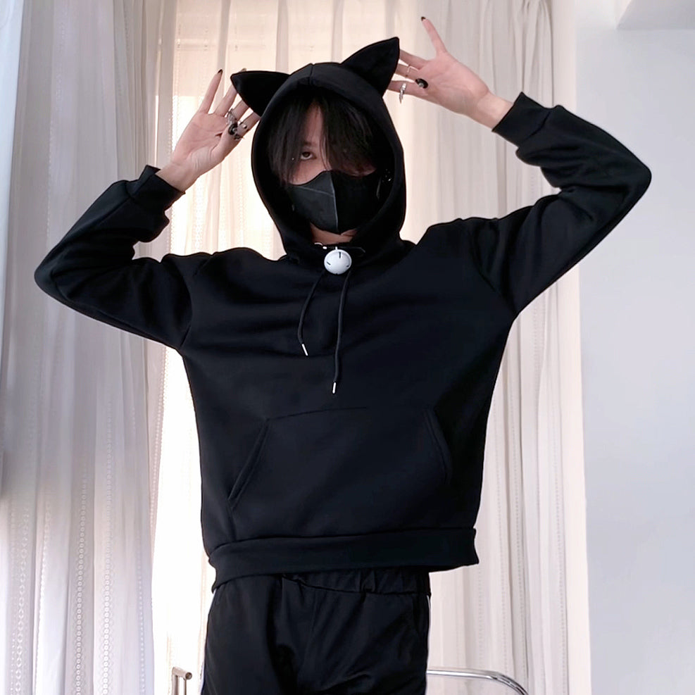 Femboy wearing a black hoodie with cat ears and a matching mask, standing against a light curtain background.