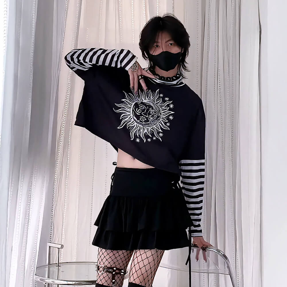 Person wearing a black cropped sweatshirt with a sun design and a black skirt, standing against a white curtain.