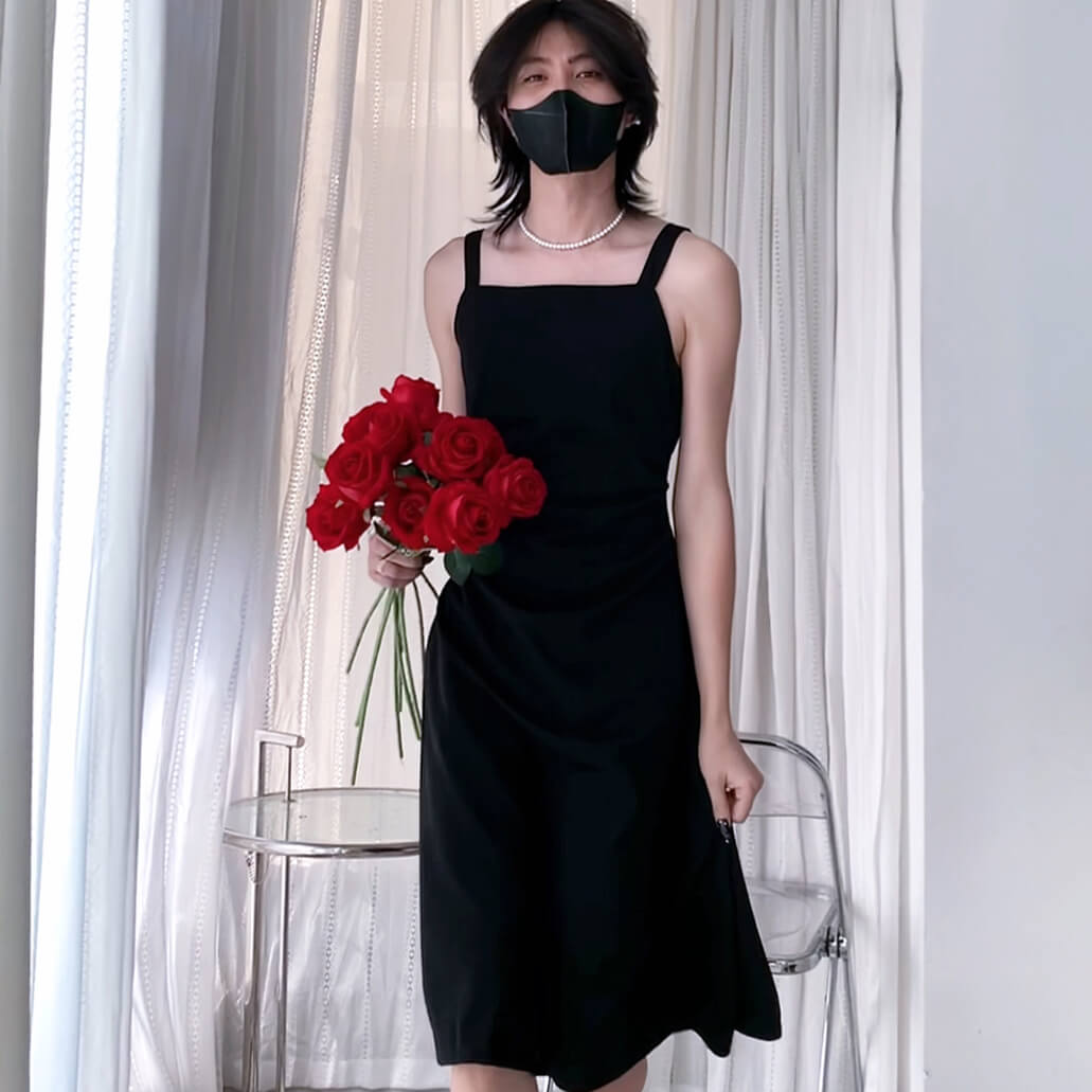 Femboy wearing a black dress and holding red roses in front of white curtains