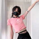 Person wearing a pink crop top with a white design and black pants, standing against a light curtain background.