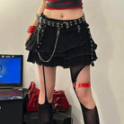 Person wearing a black tiered skirt with chains, red top, and black tights in a room with a laptop and red bag.