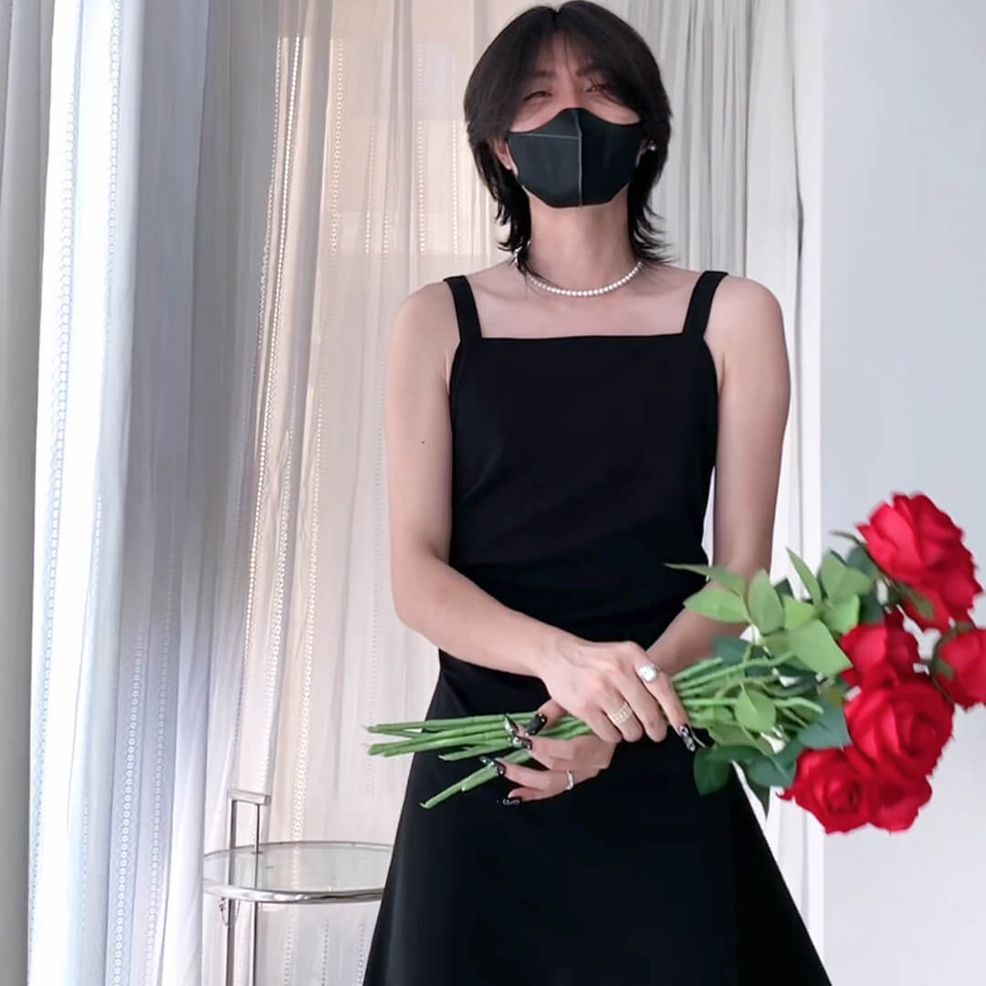 Femboy in a black dress holding red roses indoors with curtains in the background