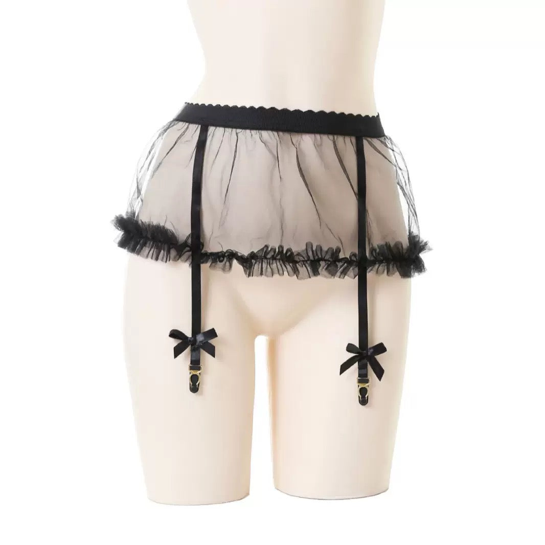 Black sheer mesh garter belt with ruffled trim and two hanging suspender straps with bow details, displayed on a beige mannequin torso against a white background.