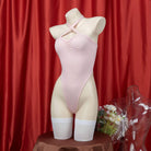 A pink halter bodysuit lingerie displayed on a mannequin against a red backdrop, with a white flower arrangement in front.