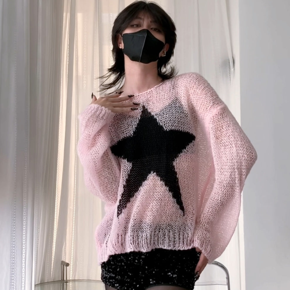 A femboy wearing a light pink chunky knit sweater with a large black star graphic on the front, paired with a black face mask and black sequined bottoms, standing in front of white curtains.