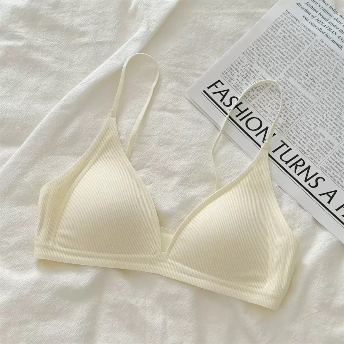 A cream-colored ribbed triangle bralette with thin straps, laid flat on a white textured fabric next to a printed magazine page.