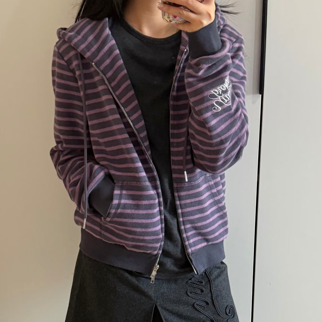 A femboy wearing a zip-up hoodie with horizontal purple and dark gray stripes, layered over a black shirt and dark skirt, taking a mirror selfie.