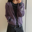 A femboy wearing a zip-up hoodie with horizontal purple and dark gray stripes, layered over a black shirt and dark skirt, taking a mirror selfie.