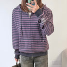 A femboy takes a mirror selfie wearing a purple and pink striped zip-up hoodie with dark gray ribbed cuffs, hem, and drawstrings, paired with gray jeans.