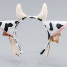 A plush cow-themed headband with white faux fur, black spots, two cream horns, pink inner lining, and ribbon bows on a clear acrylic stand against a light gray background.