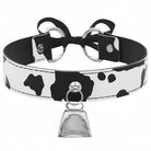 A black and white cow-print faux leather choker collar with silver-tone metal hardware, featuring a bow at the back and a dangling bell charm.