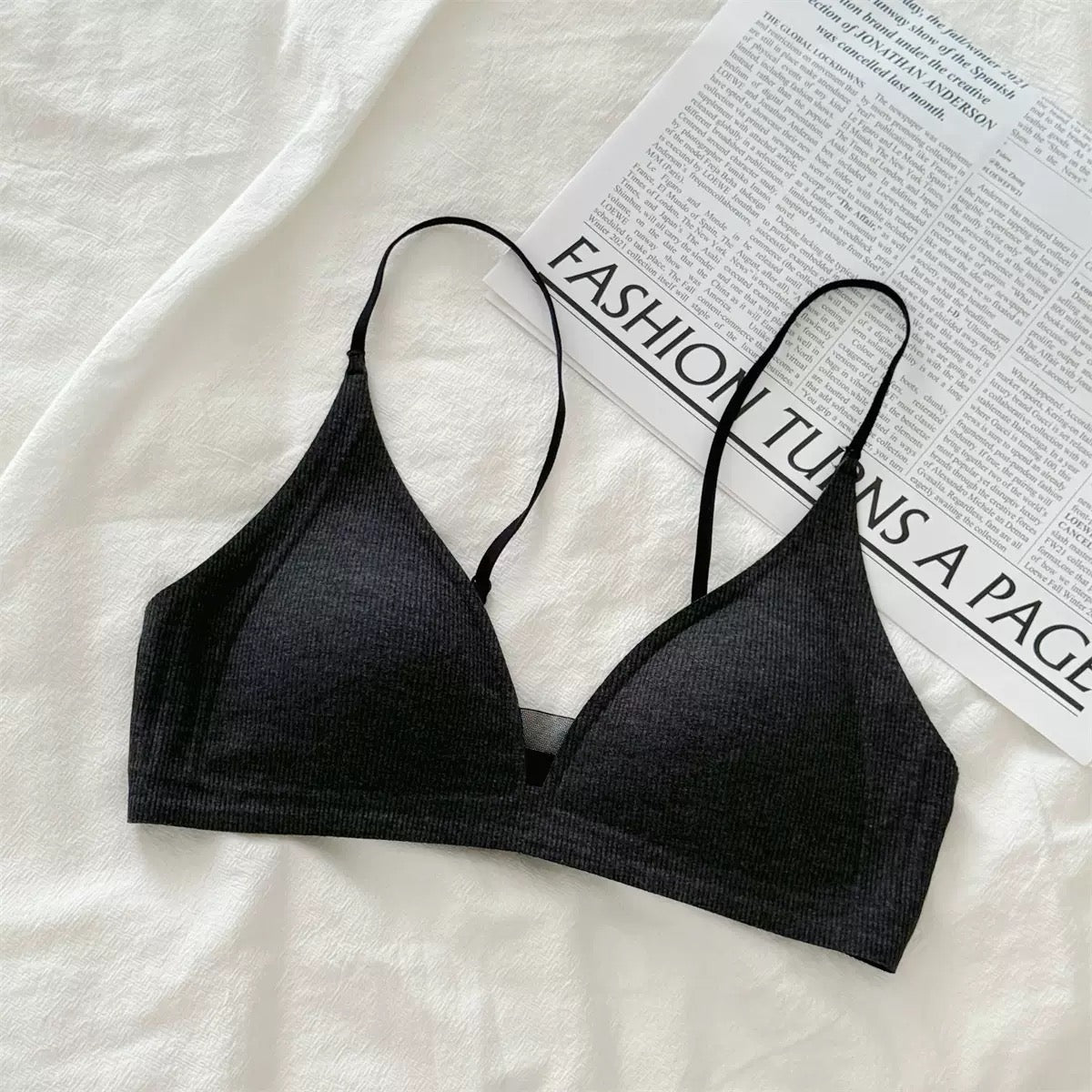 A black ribbed knit triangle bralette with thin adjustable straps, laid flat on a white textured fabric surface next to a printed magazine page.