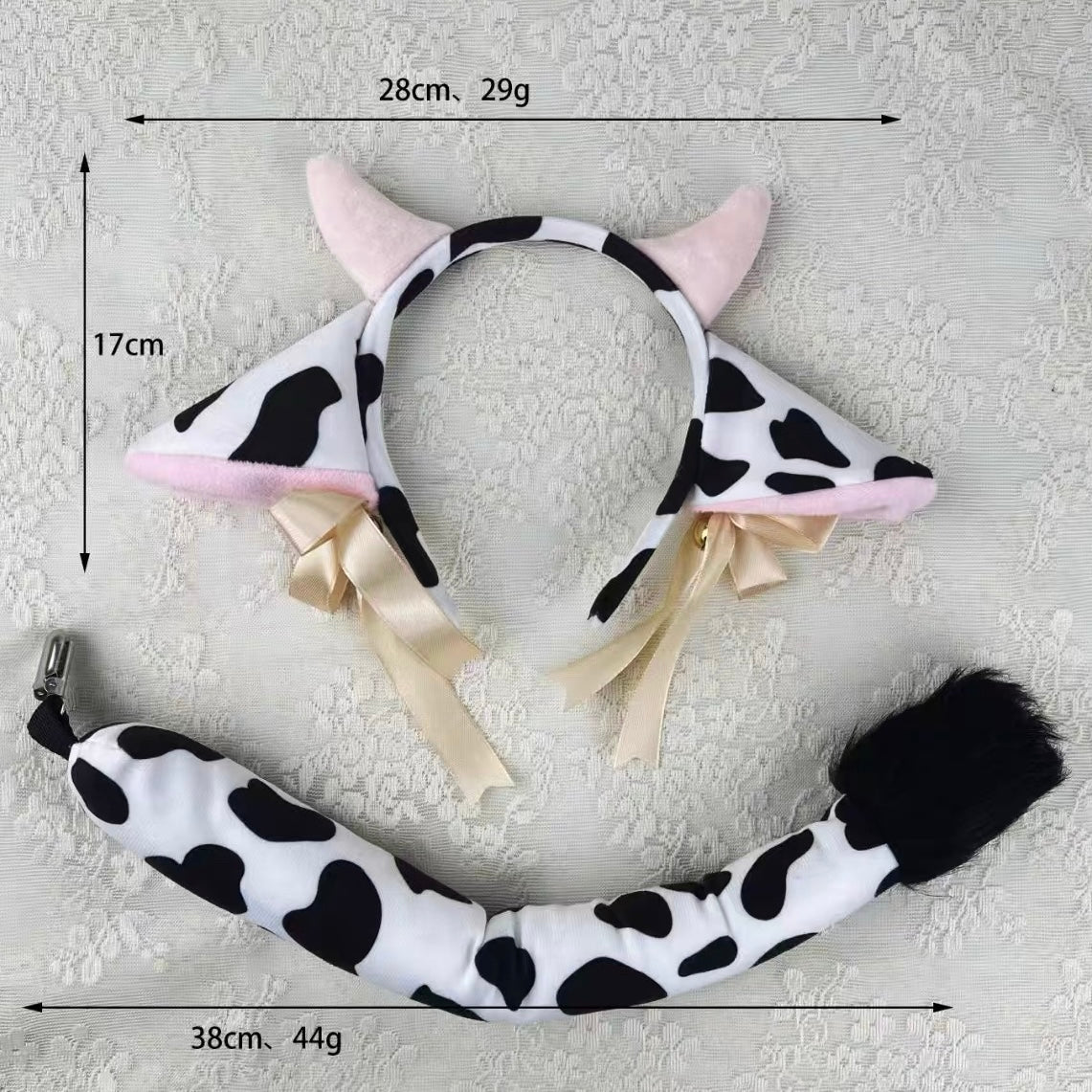Cow-print plush headband with pink horns, cream ribbons, and a matching tail accessory laid on a light textured fabric background, with measurement lines and text showing dimensions and weight.