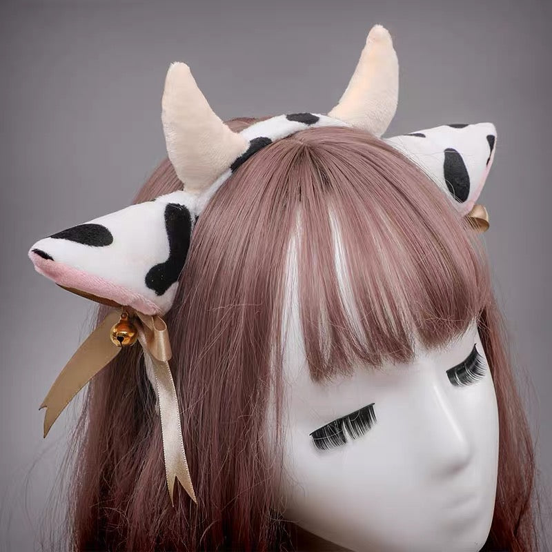 A cow-themed headband with plush ears, black spots, cream horns, and a gold ribbon, shown on a mannequin head with pink-brown hair against a gray background.