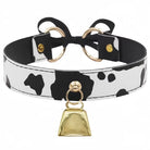 A cow-print faux leather collar with black and white spots, featuring a black bow at the back, gold-tone metal rings, and a hanging gold bell.