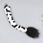 A black and white cow-print faux fur tail accessory with a black faux fur tip and a silver metal clip, displayed on a light gray background.