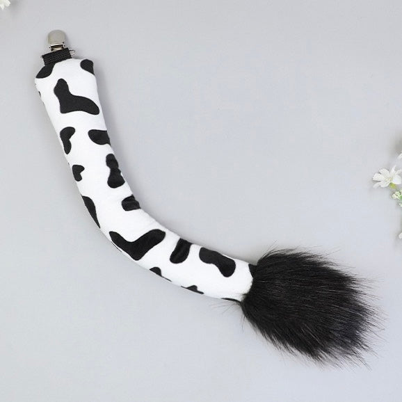 A black and white cow-print faux fur tail accessory with a black faux fur tip and a silver metal clip, displayed on a light gray background.
