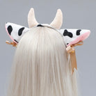 A white faux fur cow headband with black spots, pink inner ears, and cream horns, shown on a model with long white hair from the back.