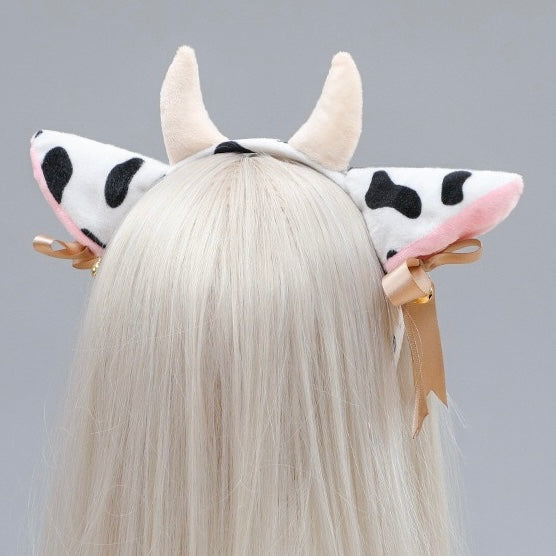 A white faux fur cow headband with black spots, pink inner ears, and cream horns, shown on a model with long white hair from the back.