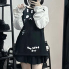 A femboy takes a mirror selfie wearing a black sleeveless pinafore-style dress with white cartoon graphics and text, layered over a white long-sleeve top. The setting appears to be a boutique or room with shelves and shoes visible in the background.
