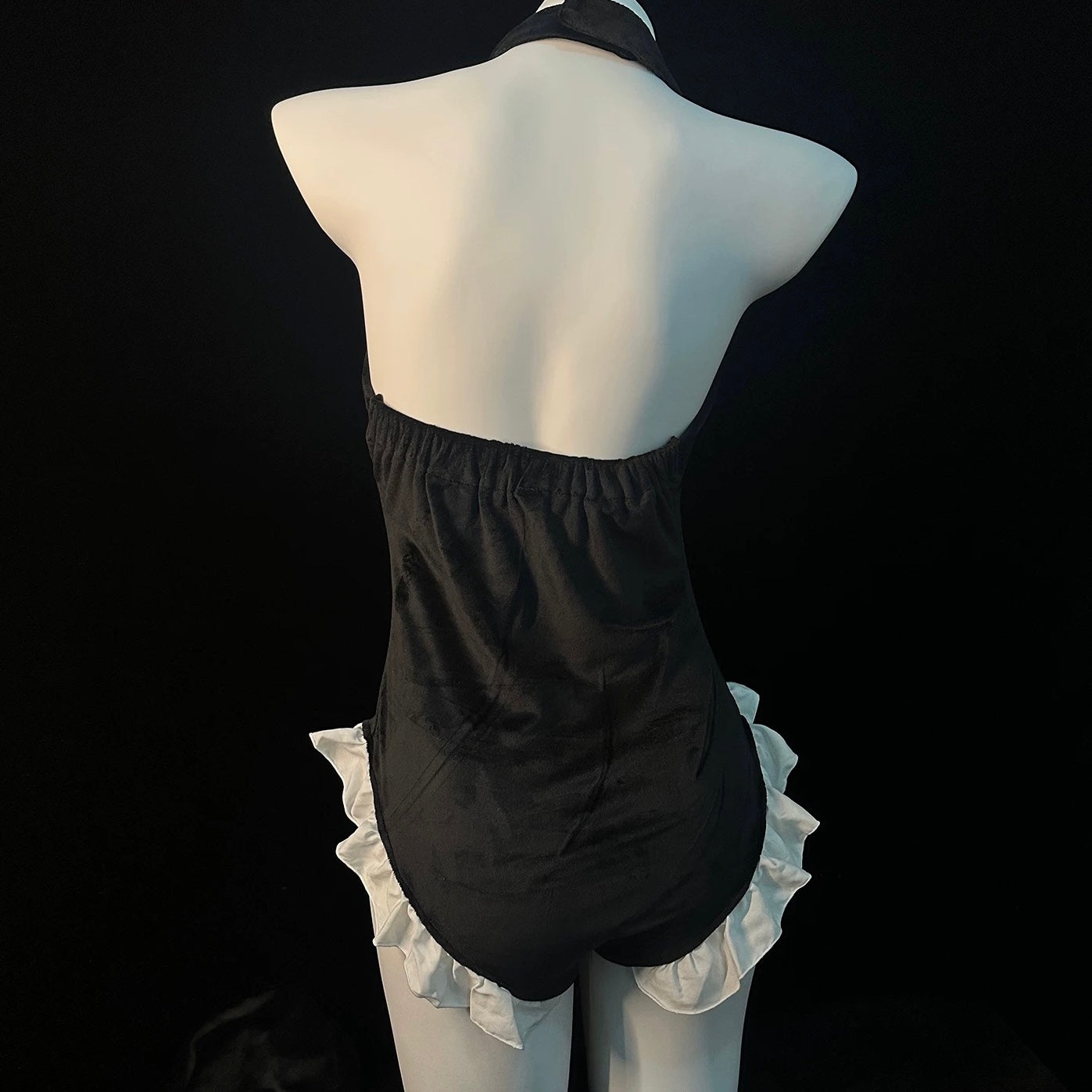 Black halter bodysuit with white ruffled trim, displayed on a white mannequin against a black background.