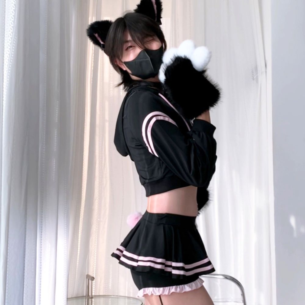 A femboy wearing a black cropped hoodie and pleated mini skirt set with pink and white stripes, black cat ears, and a black-and-white fluffy tail accessory, posed indoors against white curtains.
