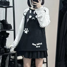 A femboy taking a mirror selfie in a clothing store wearing a black and white outfit. The outfit consists of a black pinafore-style dress with white graphic prints (cat face, paw print, and text) layered over a white long-sleeve shirt, and a black pleated mini skirt.