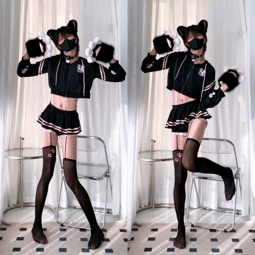 A femboy wearing a black  crop hoodie and mico skirt set, holding a black plush toy in a room setting.