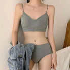 A femboy wearing a ribbed gray two-piece lingerie set consisting of a V-neck camisole top and matching high-waisted briefs, with a light blue denim jacket draped over one arm, set against a neutral indoor background.