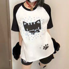 A person wearing a white and black raglan t-shirt with a black cat graphic, black shorts, and black fuzzy arm warmers, standing indoors.