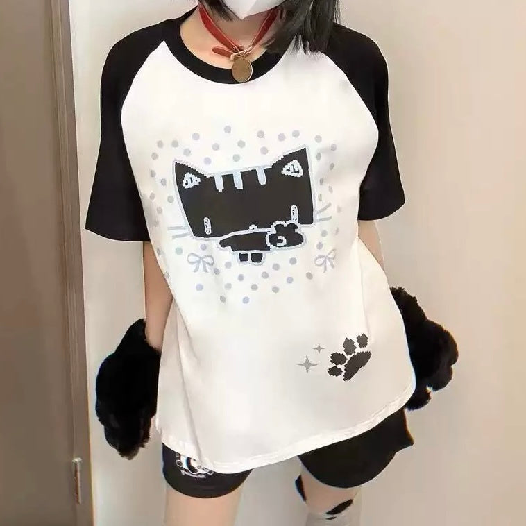 A person wearing a white and black raglan t-shirt with a black cat graphic, black shorts, and black fuzzy arm warmers, standing indoors.