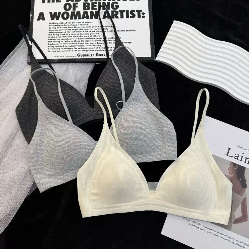 Three wireless triangle bralettes in light gray, medium gray, and cream are arranged in a flat-lay with styling props including a wavy white strap, a magazine page, and a black fabric background.