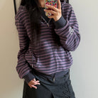 A femboy taking a mirror selfie wearing a dark purple and black horizontally striped zip-up hoodie with a white embroidered logo on the sleeve, paired with a dark gray skirt.