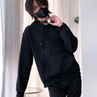 Femboy wearing a black hoodie and pants with a mask indoors.