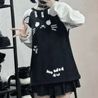 A femboy wearing a black cat-face print mini dress over a white long-sleeve shirt, standing in front of a mirror with shoes on a shelf in the background.