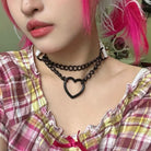 Person wearing a black chain necklace with a heart pendant, pink hair, and plaid shirt.