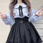 Person wearing a blue blouse with ruffled collar and long sleeves, black skirt with heart-shaped belt, standing against a light-colored wall.