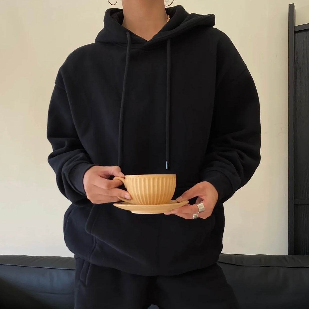Person wearing a black hoodie holding a beige cup and saucer.